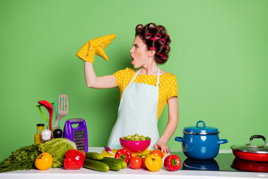 Profile side photo of shocked girl prepare tasty supper dish talk speak pot holder wear dotted yellow t-shirt hair rollers table cucumber tomato ingredients isolated green color background