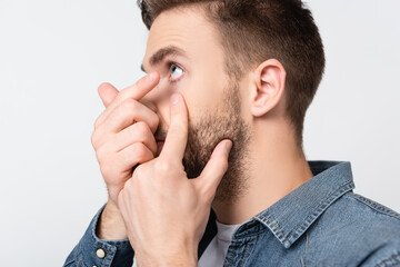 Young man wearing contact lens and touching cheek isolated on grey