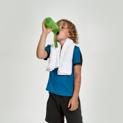 Little sportive boy child in sportswear drinking water from the bottle while standing with towel around his neck isolated over white background