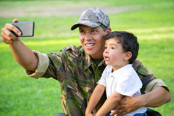 Cheerful disabled military dad and his little son taking selfie together in park. Boy sitting on...