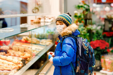Kid boy wearing medical mask buy bread and pastry for school lunch in bakery. Child with backpack and winter clothes. Schoolkid during lockdown and quarantine time during corona pandemic disease
