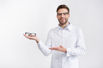 Front view of happy doctor holding and pointing with hand at eyeglasses on palm, while looking at camera isolated on white