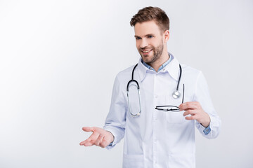 Smiling young adult doctor holding eyeglasses, gesturing, while looking away isolated on white