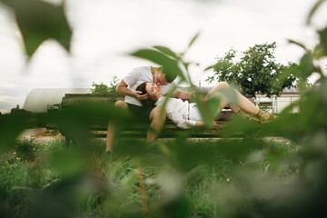 Young couple enjoying time together - woman lying on knees of man on bench on summer sunny day. Smiling man and woman are kissing