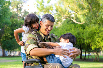 Happy disabled military dad walking with two children in park. Girl holding wheelchair handles, boy resting on dads lap. Veteran of war or disability concept