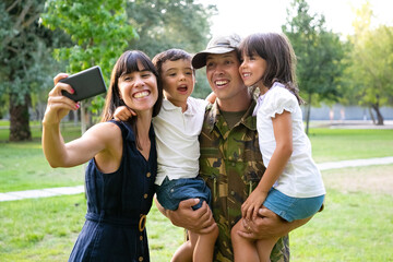Fototapeta premium Happy excited military man, his wife and two kids celebrating dads returning, enjoying leisure time in park, taking selfie on cell phone. Medium shot. Family reunion or returning home concept