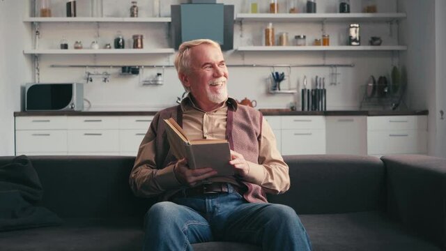 Grey haired man laughing while reading book with kitchen on background