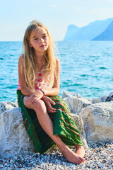 Child girl sitting on a stone on the beach. In the background lake Garda (Lago di Garda) in italy
