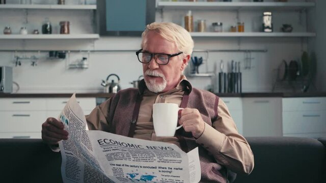 Senior man in eyeglasses reading newspaper, while drinking cup of tea at home