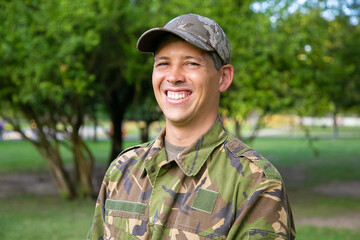 Portrait of happy man in military camouflage uniform standing in park, looking at camera and smiling. Green trees in background. Military man or guard concept
