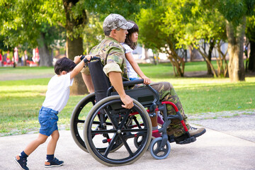 Disabled retired military man walking with children in park. Girl sitting on dads lap, boy pushing wheelchair. Veteran of war or disability concept