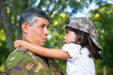 Positive dad in camouflage uniform holding offended daughter in arms, hugging girl outdoors after returning from military mission trip. Closeup shot. Family reunion or returning home concept