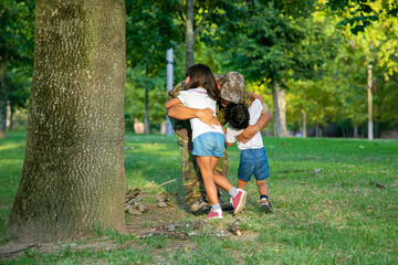 Fototapeta premium Dad meeting with two children after military mission trip, hugging kids on grass in park. Sunshine background. Family reunion or returning home concept