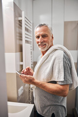 Joyous senior gentleman with a smartphone near a bathroom sink