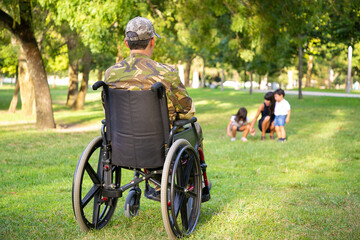 Lonely disabled retired military man in wheelchair looking at his wife and little children playing...