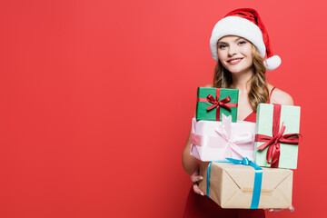 cheerful woman in santa hat holding gift boxes on red