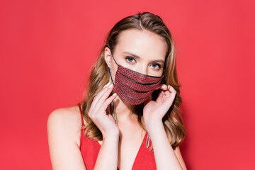 young woman wearing protective mask with rhinestones and looking at camera on red