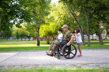 Handicapped military veteran walking with two children in park. Boy sitting on dads lap, girl...