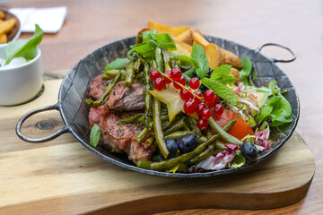 Roasted steak with green beans, mixed salad with fruits and potato wedges on an iron cooking pan on a wooden board and table, selected focus