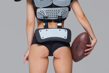 Young sporty brunette wearing sexy uniform of American football player posing with ball looking at camera isolated on grey background