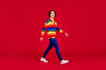 Fototapeta na wymiar Full length profile photo of beautiful attractive young lady good mood walking street friends meeting wear rainbow jumper cardigan trousers shoes isolated bright red color background