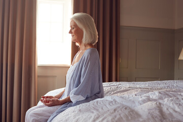 Senior Woman Sitting On Edge Of Bed At Home Meditating During Lockdown For Covid-19