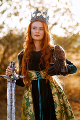 Queen with red hair in a green dress with a crown and a sword near a horse with a bird