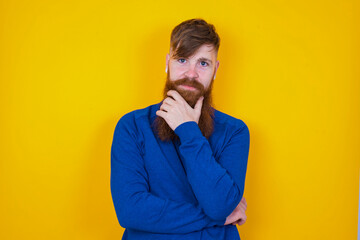Thoughtful smiling Young handsome red haired bearded man standing against yellow wall,  keeps hand under chin, looks directly at camera, listens something with interest. Youth concept.