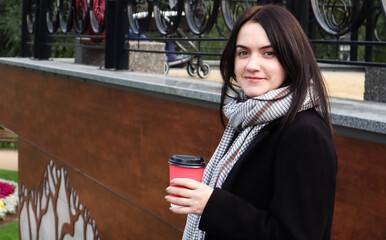 Young stylish woman in a coat and scarf drinks morning hot coffee in a red eco paper glass outdoors...