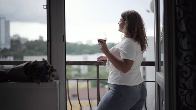 Side view of happy young chubby woman enjoying overcast evening on balcony. Portrait of relaxed Caucasian lady drinking wine from glass and smiling. Positivity and lifestyle.