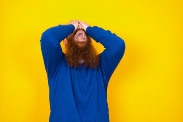 Shocked Young handsome red haired bearded man standing against yellow wall holding hands on face...