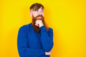 Face expressions and emotions. Thoughtful Young handsome red haired bearded man standing against yellow wall holding hand under his head, having doubtful look.