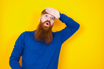 Young handsome red haired bearded man standing against yellow wall wiping forehead with hand making...