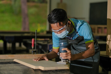 Fototapeta na wymiar Young carpenter in work clothes and face mask using trimmer for console table