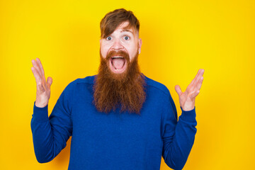 Joyful excited lucky Young handsome red haired bearded man standing against yellow wall cheering, celebrating success, screaming yes with clenched fists