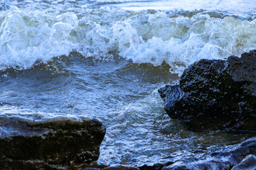 Fototapeta premium waves run onto the shore and crash against the rocks, creating many splashes and splashes near the shore. river surf in stormy weather near a stone pebble coast with foamy splashing waves.