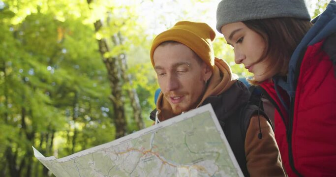 Close up portrait of loving couple handsome man and woman sitting in woods on stump and looking, examining studying map, discussing a route plan, man showing direction with hand. Journey in Wildlife