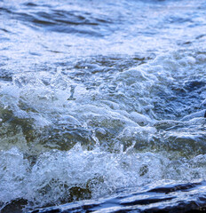 Obraz premium waves run onto the shore and crash against the rocks, creating many splashes and splashes near the shore. river surf in stormy weather near a stone pebble coast with foamy splashing waves.