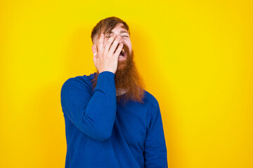 Charismatic carefree joyful Young handsome red haired bearded man standing against yellow wall likes laugh out loud not hiding emotions giggling hear funny hilarious joke chuckling facepalm.