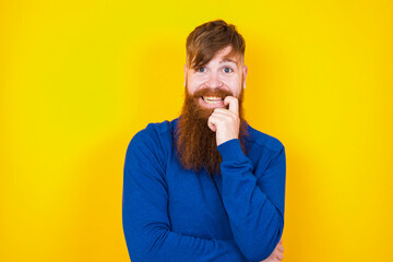 Carefree successful Young handsome red haired bearded man standing against yellow wall touching jawline gazing camera tilting head grinning white teeth delighted. Dental care concept.