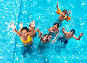 Boys with girls jump and play in swimming pool, splashing, lift hands smile view from above