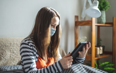 Young woman in a protective mask is sitting at home on the bed and using her tablet. Concept of online communication, remote work and lifestyle in the pandemic