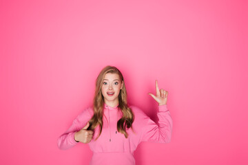 Obraz na płótnie Canvas smiling beautiful woman pointing with finger and showing thumb up on pink background