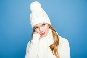 pensive blonde beautiful woman in winter white outfit on blue background