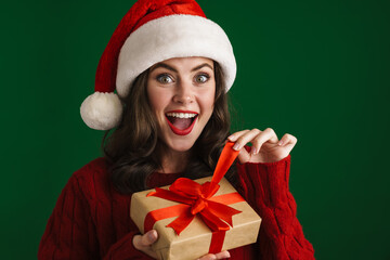 Charming delighted girl in Santa Claus hat opening Christmas gift