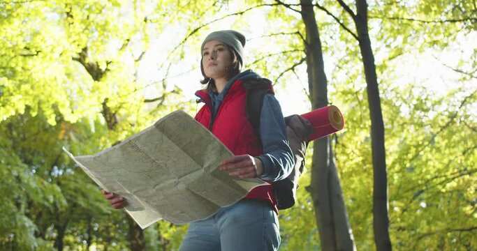Close up of woman standing in forest, examining route plan. Female lost in wood and searching way back. Tourist looking at map while hiking with trekking poles. Traveling in nature.