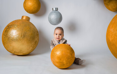 a beautiful little boy in a suit with a bow tie sits with a Golden ball on a white background with giant holiday balloons