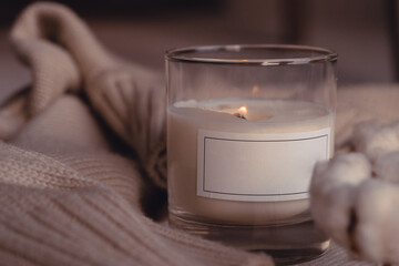 Symbol Of Coziness. Burnng Scented Aroma Candle In Glass With Blank White Label Near Cotton Flower On Cozy Stylish Sweater