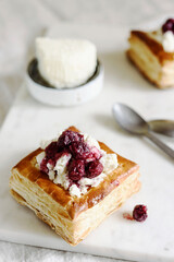 Puff pastry squares with ricotta and cherry sauce on white marble tray. Baked small square cakes topped with cheese and berry sauce