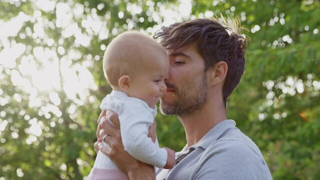 Loving father cuddling and kissing baby daughter as he holds her outdoors in park or garden against flaring sun - shot in slow motion